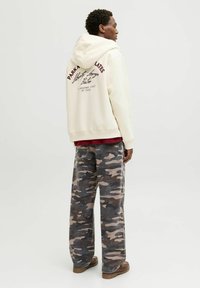Un sweat à capuche crème clair avec du texte bordeaux, associé à un pantalon camouflé gris ample. Des chaussures marron robustes complètent cette tenue décontractée.