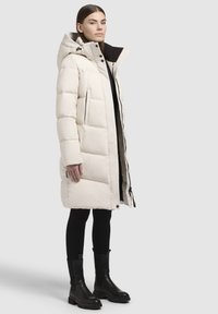 Lange witte pufferjas met een capuchon, gestikte textuur, dubbele zijzakken en een zwarte binnenvoering, gecombineerd met zwarte enkelboots en leggings.
