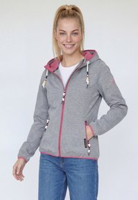 Jeune femme souriante, portant un sweat à capuche gris zippé avec doublure rose et un jean bleu, une main dans la poche, debout devant un fond uni.