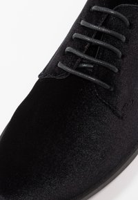 Chaussure de ville en velours noir avec surface lisse, bout arrondi et cinq lacets noirs. Détails de couture subtils près de la semelle.