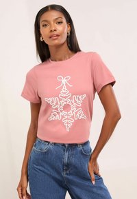 Camiseta de manga corta en color rosa con un diseño de copo de nieve y lazo en blanco en la parte frontal. Confeccionada en una tela suave, con un corte relajado y un escote tipo crew.
