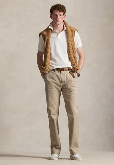 Άνδρας στέκεται με τα χέρια στις τσέπες, φοράει λευκό πόλο, μπεζ παντελόνι, πλεκτή ζώνη, λευκά αθλητικά παπούτσια και καφέ πουλόβερ περασμένο στους ώμους.