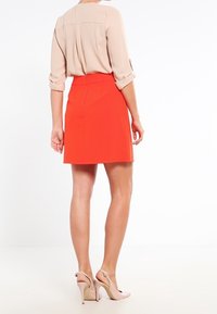 Červená áčková sukně s vysokým pasem, hladká tkanina, zkombinována se světlou béžovou halenou s dlouhým rukávem a nahými sandály na vysokém podpatku.