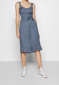Un vestido azul con lunares que presenta un cuerpo ajustado, botones en la parte delantera y un cinturón a la cintura, con una falda acampanada y un dobladillo con volantes, combinado con zapatillas blancas.