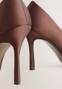 Brune satin høje hæle med spids tå og slank silhuet. Har en glat tekstur og en sort detalje på hælen.