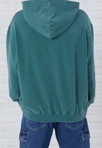 Felpa con cappuccio color turchese, caratterizzata da una texture morbida, polsini e orlo a coste, con vestibilità rilassata, indossata con pantaloni cargo blu su uno sfondo bianco.
