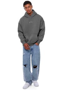 Grauer Hoodie mit einer Vordertasche und einem Logo-Patch, kombiniert mit hellblauen, ausgewaschenen, weiten Jeans mit Rissen, abgerundet mit weißen Sneakers.