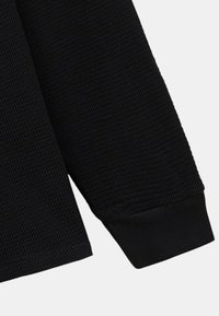 Vêtement noir à manches longues en tissu texturé et côtelé. Présente des poignets ajustés et un col droit. Design minimaliste sans matériel visible.