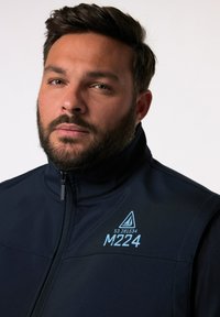 Navy Zip-Up-Jacke aus glattem Stoff; verfügt über einen hohen Kragen und ein gedrucktes, hellblaues Logo auf der linken Brustseite mit einem Segelboot-Design.