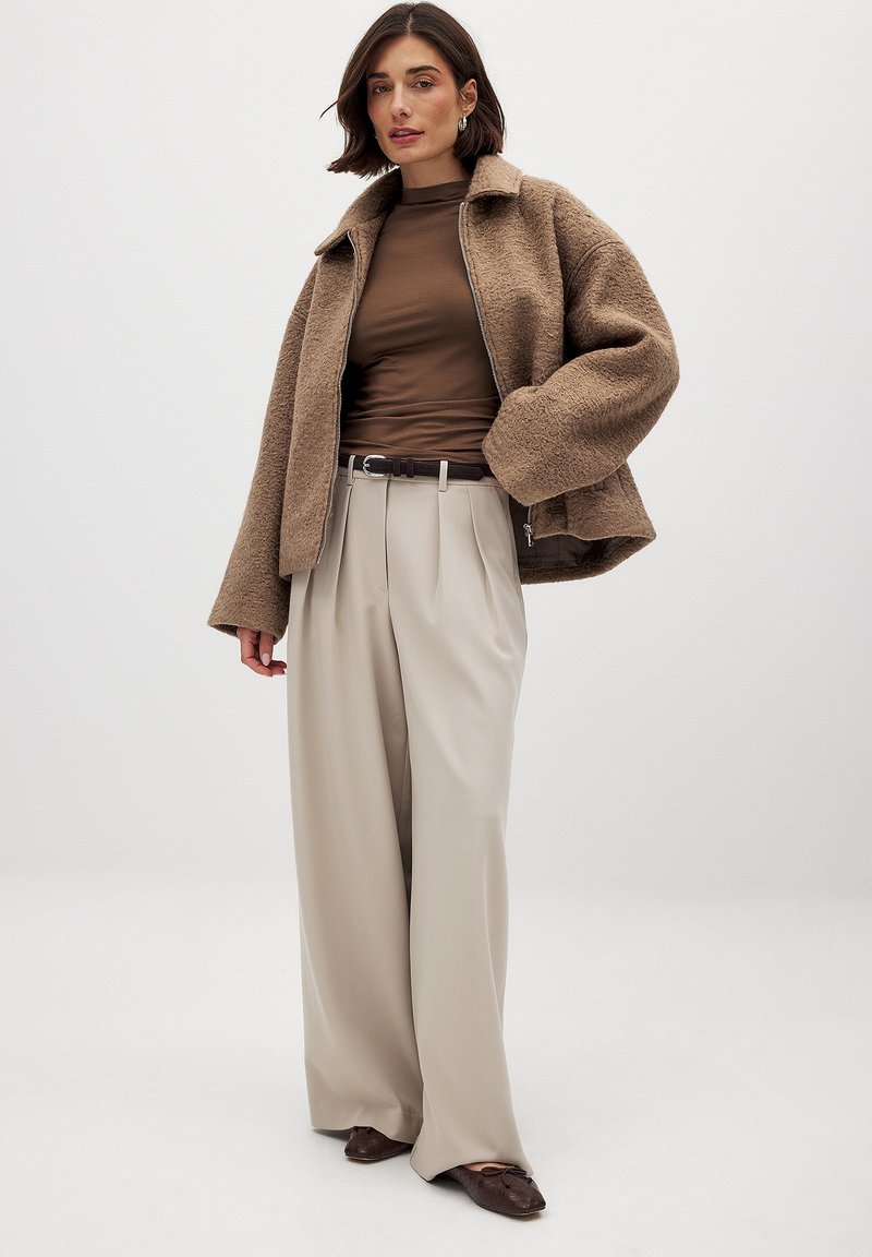 Veste brun texturée avec un col, portée avec un haut brun ajusté et un pantalon beige large, complété par une ceinture et des chaussures sombres.