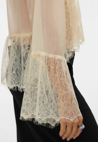 Valge beežist pluusivarrukas detailse pitsäärega mustade pükste kohal, kus on näha käsi sinise kiviga sõrmusega.