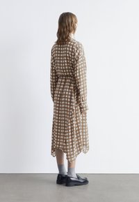 Braun- und cremefarbenes karriertes Kleid mit tailliertem Schnitt, langen Ärmeln und asymmetrischem Saum, kombiniert mit schwarzen Schuhen und grauen Socken.