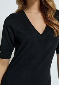 Haut en tricot noir avec un décolleté en V, des manches courtes retournées et une texture lisse, avec des détails discrets au niveau du décolleté et des manches.