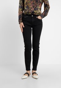 Vaqueros skinny de denim negro con una cintura de media altura, que cuentan con bolsillos frontales y cierre de botón. Combinados con una blusa de manga larga con estampado y zapatos.