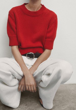 Maglione rosso in maglia con un ampio scollo a coste, maniche corte, abbinato a pantaloni bianchi larghi, accentuati da una cintura nera con fibbia d'argento.