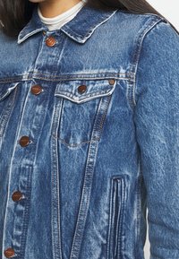 Kvinna som bär en medelmörk blå denimjacka med kopparfärgade knappar och ficka fram över en vit skjorta.