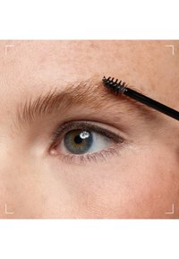 Gros plan d'un œil avec une iris bleue et un sourcil coiffé avec une brosse spoolie noire pour mascara.