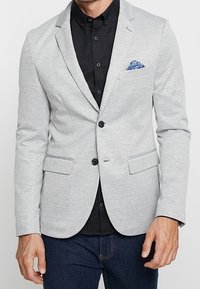 Hombre con una blazer gris claro sobre una camisa negra abotonada, con un pañuelo de bolsillo azul con estampado y vaqueros azul oscuro.