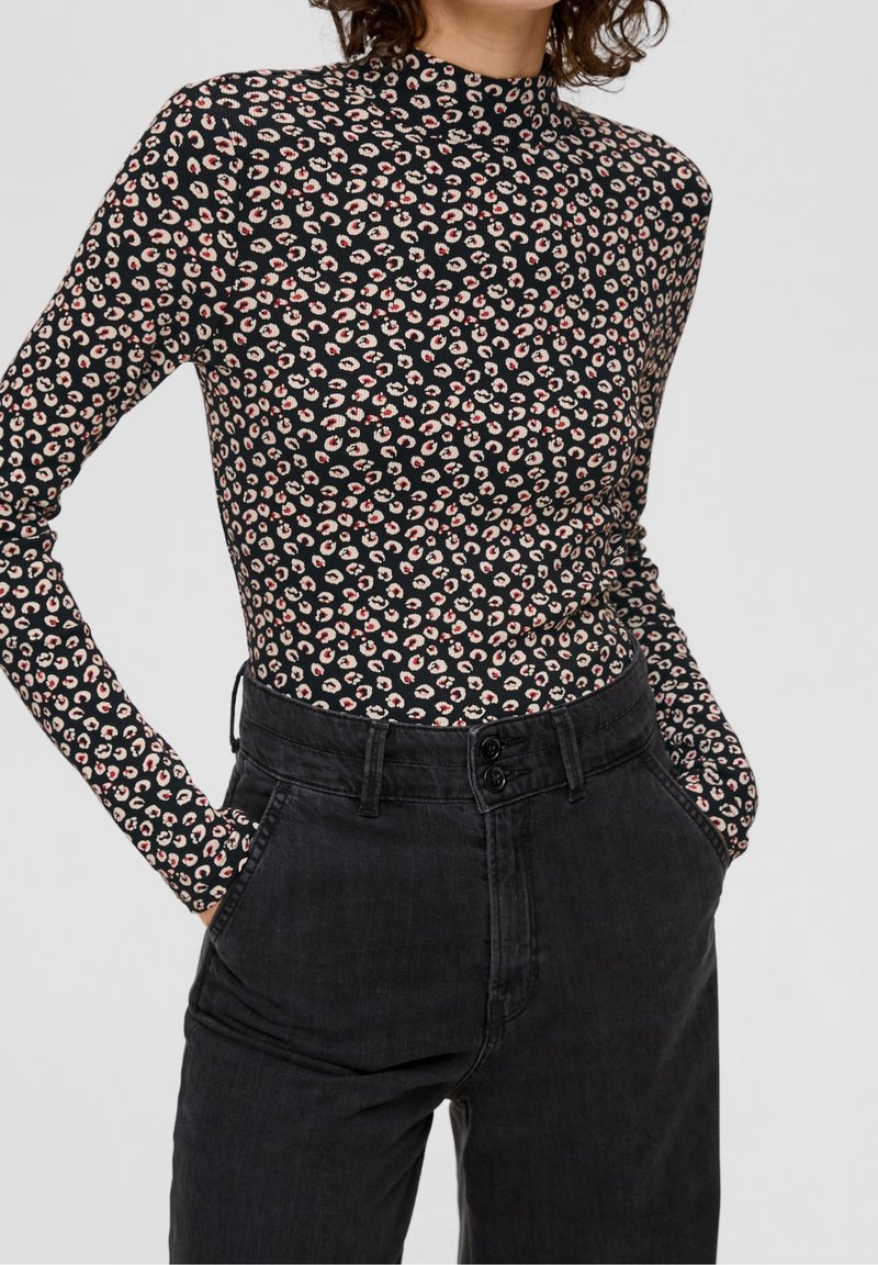 Personne portant un haut noir ajusté à manches longues avec de petites taches léopard beige et rouge, rentré dans un jean noir taille haute, les mains dans les poches.