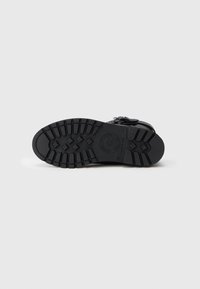 Sola de bota preta com padrão de solado profundo apresentando formas retangulares e em diamante, com uma fivela visível na lateral, colocada sobre um fundo branco.