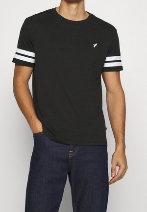 Homme portant un t-shirt noir à manches courtes avec deux bandes blanches sur chaque manche et un petit logo blanc d'oiseau sur la poitrine gauche, associé à un jean bleu foncé.