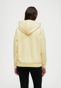 Gul hettegenser med en myk tekstur, som har en stor hette og ribbede mansjetter. Designet inkluderer en kengurulomme foran.