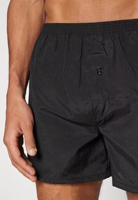Schwarze Boxer-Schlüpfer aus leichtem Stoff, mit einem elastischen Bund und einem Knopf vorne zum Schließen. Glatte Textur.
