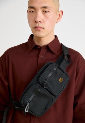 Svart stoff skulderveske med to glidelåslommer, justerbar stropp og en liten logo-patch. Båret over en burdeos skjorte.