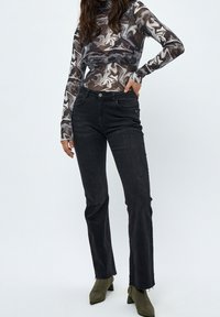 Schwarze, ausgestellte Jeans mit einem rohen Saum, kombiniert mit einem figurbetonten, mehrfarbigen Oberteil mit einem Muster aus Grautönen, Braun und Weiß.