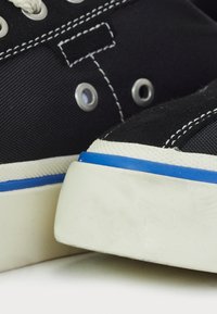 Black canvas sneakers with white stitching, metal eyelets, and a white sole featuring a blue accent stripe along the edge.