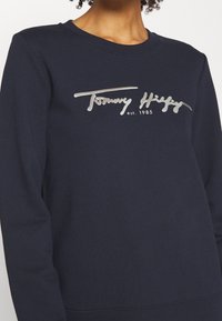 Námořnická modrá mikina s potiskem stříbrného loga "Tommy Hilfiger" a rokem "est. 1985." Měkká tkanina s klasickým kulatým výstřihem.