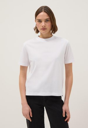 Jeune femme aux cheveux bruns courts portant un t-shirt blanc uni à manches courtes et un pantalon noir, debout devant un fond neutre.