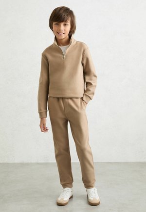 Pruunide juustega poiss, kes kannab beežikat lukuga pulloverit, sobivaid beeže pükse ja valgeid tenniseid, seisab valge seina vastu.