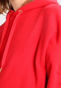 Sudadera roja con capucha ajustable mediante cordón con ojales dorados, textura suave y detalle bordado en la manga izquierda.