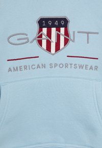 Sudadera azul claro con un logo de escudo bordado que presenta franjas rojas y blancas, "GANT" en gris y el texto "AMERICAN SPORTSWEAR".