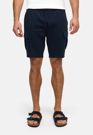Mężczyzna w granatowych krótkich spodenkach cargo z regulowanym sznurkiem w talii i czarnych sandałach z klamrą, stoi na tle białego tła.