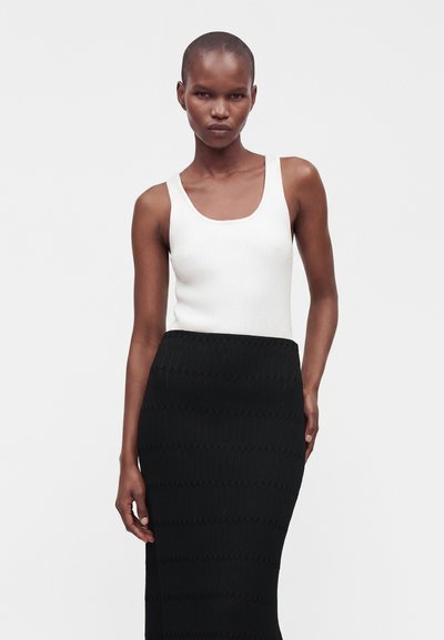 Femme aux cheveux courts portant un haut côtelé blanc sans manches et une jupe noire texturée taille haute sur un fond uni.