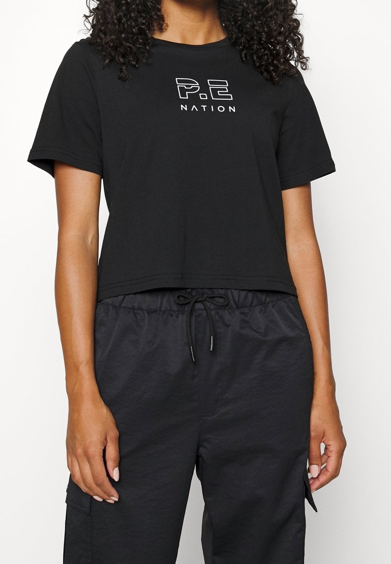 Black cropped t-shirt made of soft cotton, featuring a white logo reading "P.E NATION" on the front, paired with elastic waist, black pants.