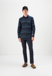 Tengerészcsíkos gombos ing, pamut anyagból, mellzsebbel rendelkezik. Sötét nadrággal és barna velúr slip-on cipővel párosítva. Fekete sapka viselve.