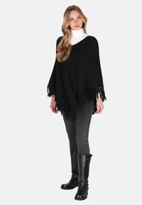 Frau mit langen, welligen Haaren, die einen weißen Rollkragenpullover, einen schwarzen poncho mit Fransen, graue Skinny-Jeans und schwarze kniehohe Stiefel trägt.