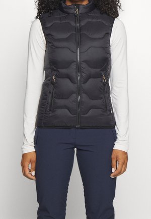 Kvinde iført en sort quiltet vest med lynlås over en hvid langærmet skjorte og marineblå bukser, stående foran en ensfarvet baggrund.