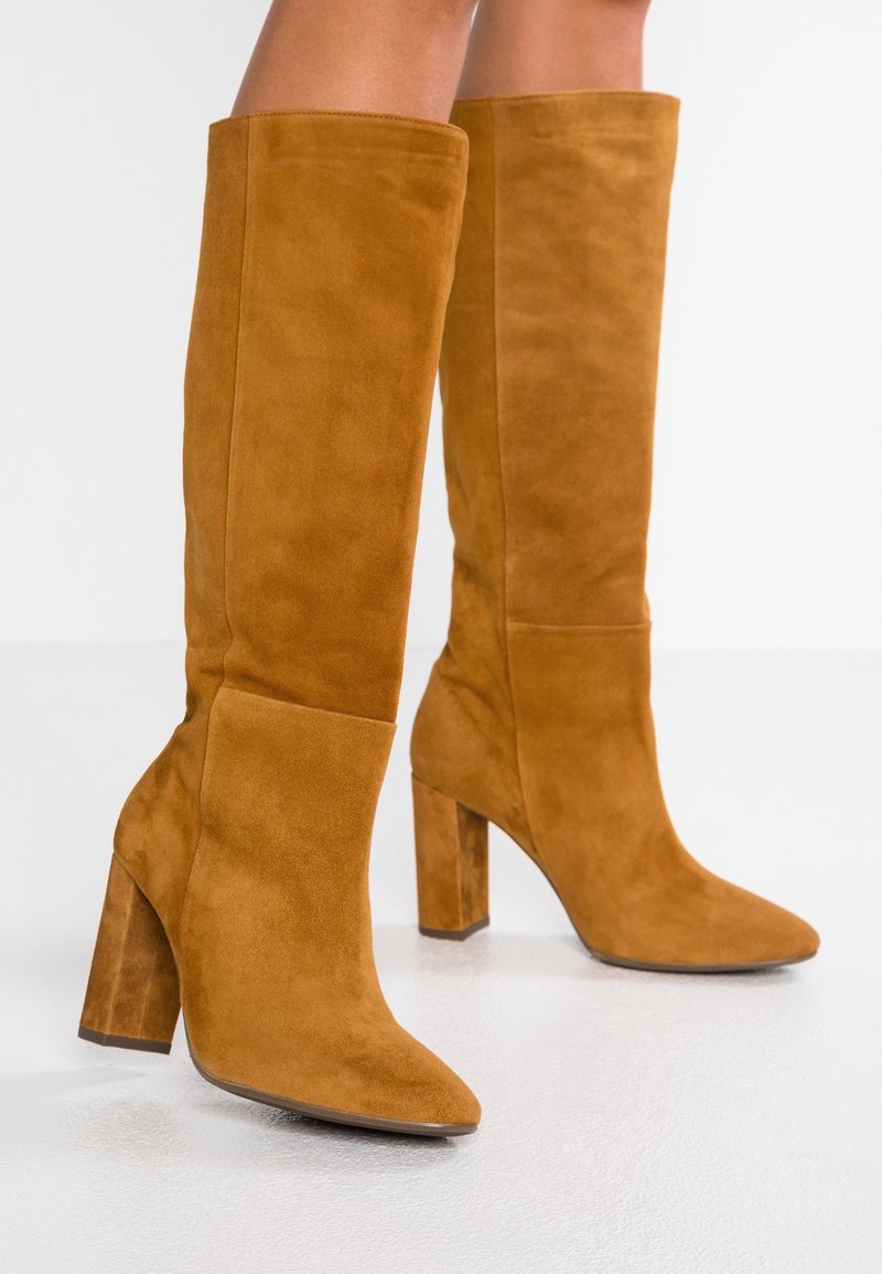 Botas altas de ante para mujer en color marrón con tacones gruesos, usadas sobre piernas desnudas contra un fondo blanco.