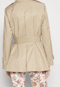 Světlý béžový trench kabát s úzkým pasem, plisovaným zadním dílem a dlouhými rukávy, doplněný květinovým vzorem na kalhotách. Přizpůsobený design a hladká textura.