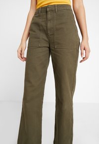 Pantaloni a gamba larga verde oliva in cotone. Caratterizzati da tasche anteriori, vita alta e un tessuto liscio e testurizzato.