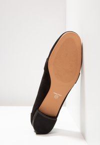 Chaussure plate noire inclinée contre un mur blanc, montrant une semelle marron lisse avec des motifs ondulés antidérapants et l'indication de taille 37.