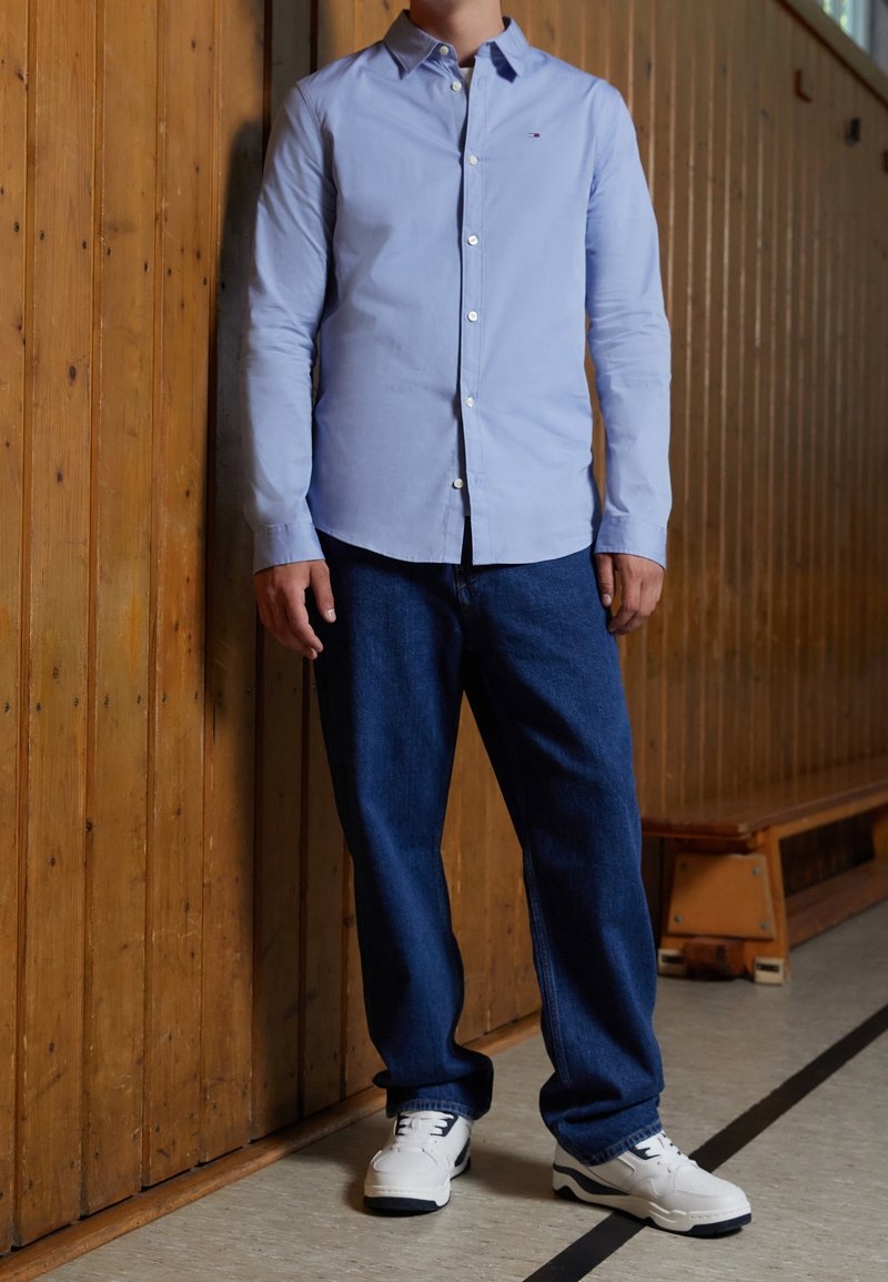 Persona che indossa una camicia azzurro chiaro con bottoni, jeans blu scuro e sneakers bianche, in piedi contro una parete in legno all'interno.