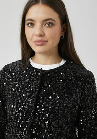 Veste noire à sequins avec un col rond et une fermeture à l'avant. Le tissu a une texture lisse, agrémentée de sequins brillants.