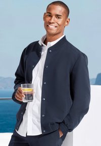 Navy blue bomber jacket with snap buttons, worn over a white shirt. Holding a glass of water with a lemon slice. Smooth fabric texture.