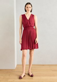 Robe sans manches bordeaux avec une taille smockée ajustée, une jupe fluide et un décolleté en V. Associée à des ballerines bordeaux assorties.
