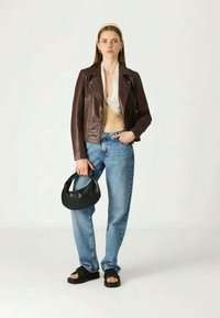 Rjava usnjena moto jakna, krema bluzica, svetlo modri kavbojke, črna torbica in črne sandale. Model stoji pred nevtralnim ozadjem.